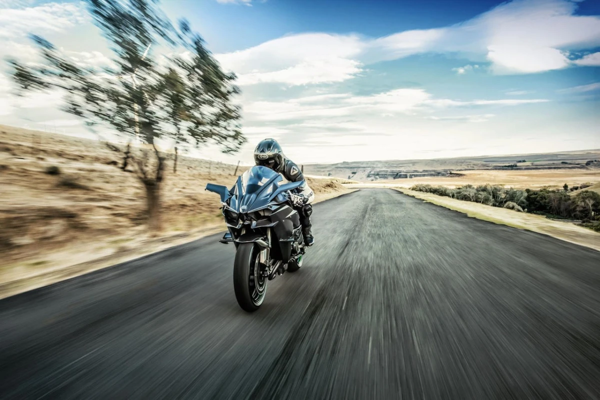 Ortaya doğru hızla gelen siyah bir Kawasaki H2R yarış motosikleti üzerinde, kasklı ve özel tulumlu bir sürücü, etrafı bulanıklaştıran yüksek bir süratle düz bir asfalt yolda ilerliyor, uzaklarda kuru tepeler ve mavi gökyüzü seçiliyor.