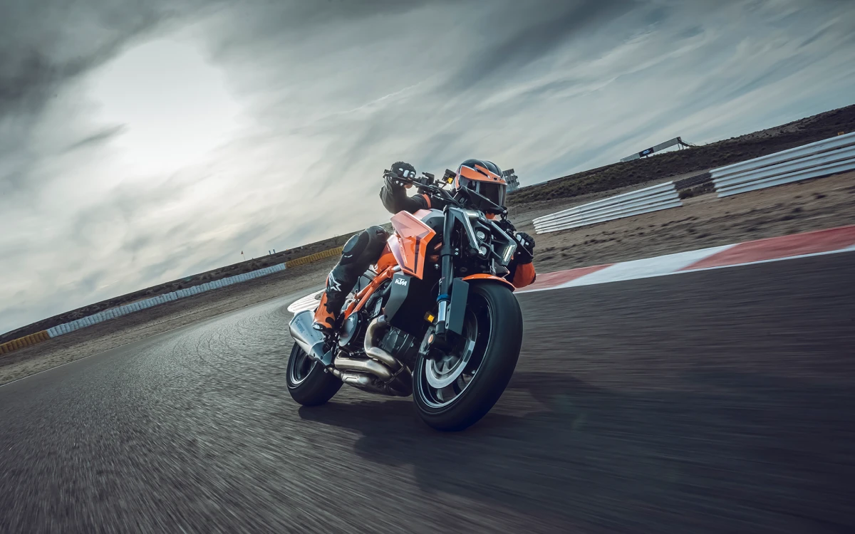 Gri bulutlu gökyüzünün altında, keskin bir virajda hızla ilerleyen turuncu ve siyah renkli bir KTM 1390 Super Duke R naked motosiklet üzerinde, tam koruyucu ekipmanlarıyla eğilmiş bir sürücü, yarış pistinin kırmızı-beyaz kerb taşları ve kenar bariyerleriyle çevrili yolunda dinamik bir an yakalanmış.