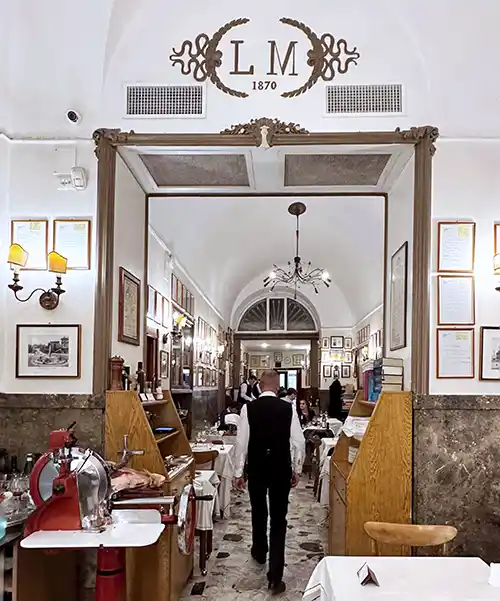 Klasik Roma restoranı La Matriciana'nın kemerli girişinden görülen, zarif beyaz örtülü masaların dizili olduğu şık iç mekanda, bir garson ileriye doğru yürürken, üstteki LM 1870 logosu bu Roma seyahat rehberinde öne çıkan mekanın köklü gastronomik tarihini gözler önüne seriyor.