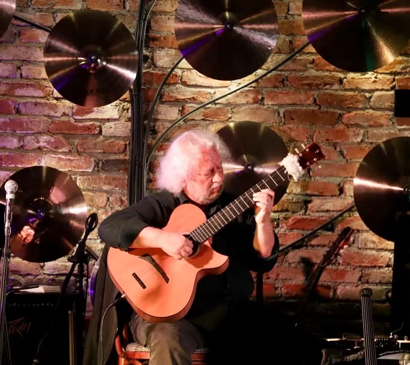 Ankara'daki Last Penny sahnesinde, beyaz saçlı ve sakallı yaşlı bir gitarist, tuğla duvarın ve arkasındaki parlak zillerin önünde telli akustik gitarını büyük bir yoğunlukla çalarken, solunda bir mikrofon standı da görünüyor.