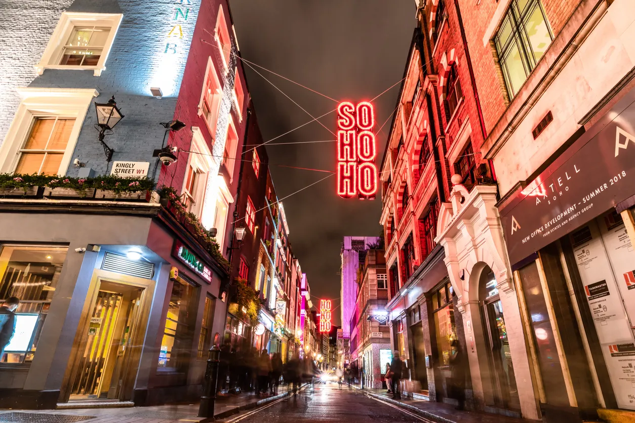 Londra'nın hareketli Soho bölgesinde, parlak neon tabelaları ve binaların ışıklarıyla aydınlatılmış, ıslak kaldırımda hareketli insan siluetlerinin yer aldığı, bir seyahat rehberi için canlı bir gece sokağı manzarası.