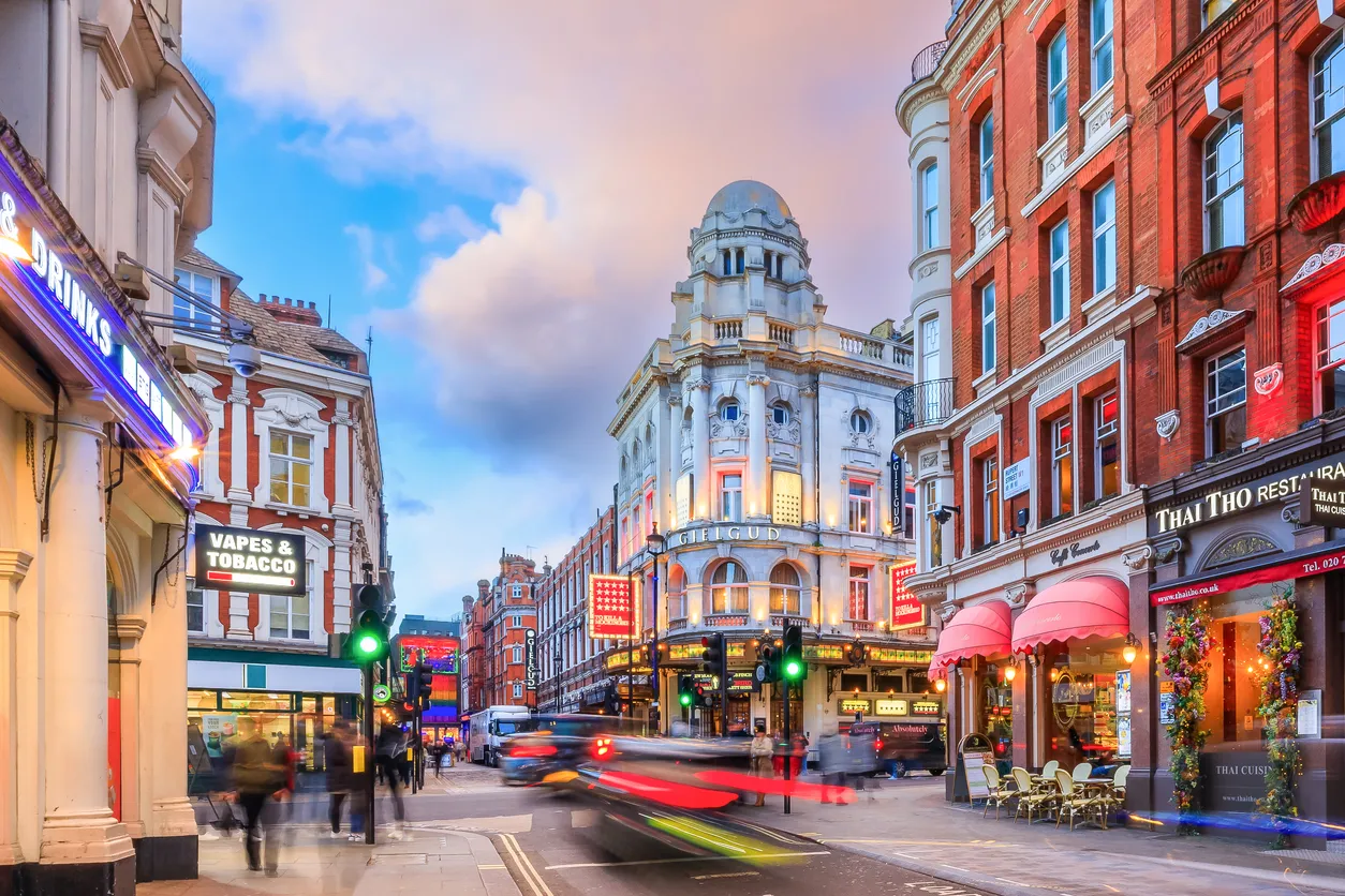 Akşamüstü ışıklarıyla aydınlanan hareketli bir Londra caddesi, tarihi binalar, neon tabelalı dükkanlar ve restoranlar, araçların oluşturduğu ışık izleriyle yoğun trafiği ve kaldırımlarda yürüyen insanları gösteren bir seyahat anı.