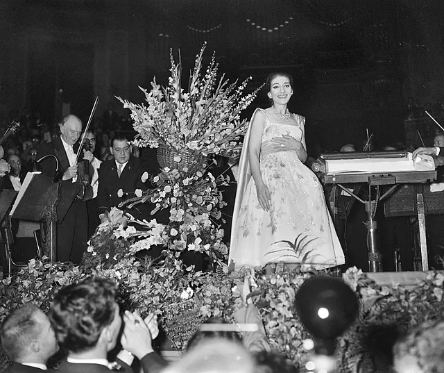 Maria Callas, büyük bir çiçek aranjmanı önünde sahnede gülümsüyor; orkestra eşliğinde alkışlarla karşılanıyor.