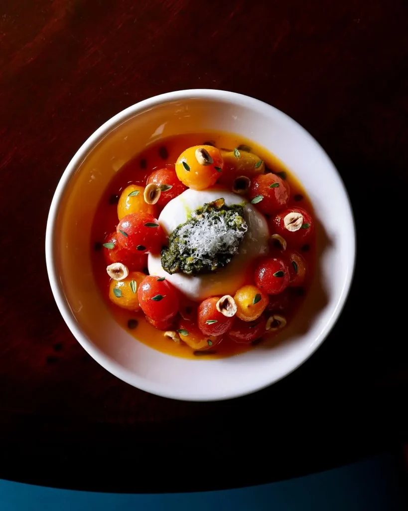 Koyu renk ahşap bir masada, ortasında kremsi burrata peyniri, etrafında kırmızı ve sarı kiraz domatesler, üzerinde pesto sosu, rendelenmiş peynir ve fındık parçacıkları ile süslenmiş bu zarif sunumlu yemek, dünyanın en iyi müze restoranlarında sunulan eşsiz lezzetlerden birini gözler önüne seriyor.