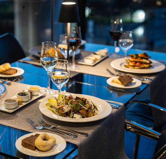 Akropolis Müzesi restoranında, akşamüstü ışıklarının vurduğu cam bir masada, çeşitli salata, ekmek ve meze tabaklarının yanında kadehlerin zarifçe dizildiği, arkada şehir ışıklarının parladığı modern ve davetkar bir müze restoranı gastronomisi deneyimi yaşanıyor.