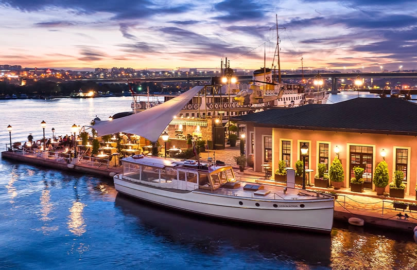 Haliç kıyısındaki Rahmi M. Koç Müzesi'nin müze restoranı Halat Restaurant'ın Haliç manzaralı iskelesi, gün batımının sıcak renkleri altında ışıklı masaları, iskeleye bağlı beyaz bir tekne ve uzakta köprü ışıklarıyla büyüleyici bir atmosfer sunuyor.