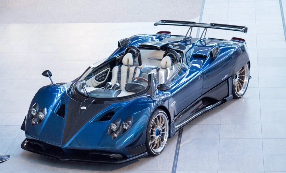 Mavi Pagani Zonda HP Barchetta hiper otomobil, V12 motoru, 789 beygir gücü ve karbon fiber detaylarıyla ışıklı bir showroomda üstten çekilmiş açıyla dikkat çekiyor, açık kokpitinde özel tasarım beyaz ve mavi ekose desenli koltuklar ve zarif el işçiliği öne çıkıyor.