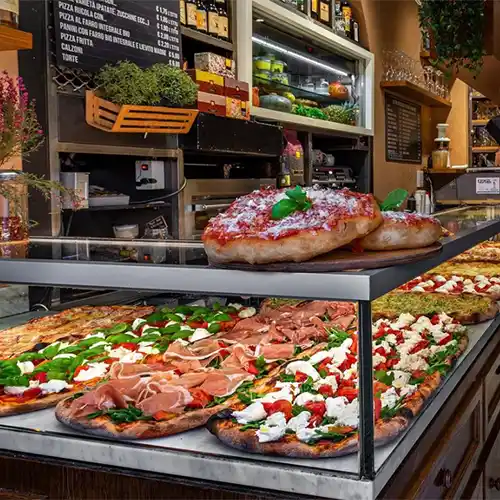 Roma Seyahat Rehberi makalesiyle uyumlu olarak, İtalya'nın başkenti Roma'da popüler bir pizzacının camekanında taze malzemelerle hazırlanmış, üzerinde mozzarella, kiraz domates, fesleğen ve proşüto gibi çeşitli lezzetlerin bulunduğu, dilimlenmeye hazır uzun dikdörtgen ve yuvarlak birçok pizza iştah açıcı bir şekilde sergileniyor.