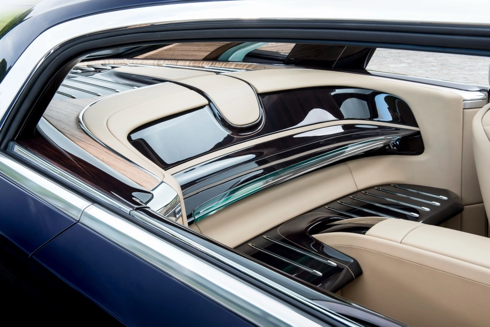 Koyu lacivert dış yüzeyli Rolls-Royce Sweptail'in arka yan penceresinden görünen, açık bej rengi deri döşemeleri, koyu ahşap kaplamaları ve parlak metal detaylarıyla göz alıcı el işçiliği ürünü lüks iç mekan detayları.