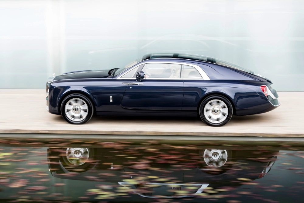 Koyu lacivert renkli, pürüzsüz hatlara sahip, kendine özgü fastback tavan tasarımına sahip lüks Rolls-Royce Sweptail otomobil, hafifçe bulanık tekerlekleriyle ilerlerken, altındaki su birikintisinde sonbahar yaprakları arasında yansıması belirginleşiyor ve dünyanın en pahalı otomobilleri listesindeki yerini vurguluyor.