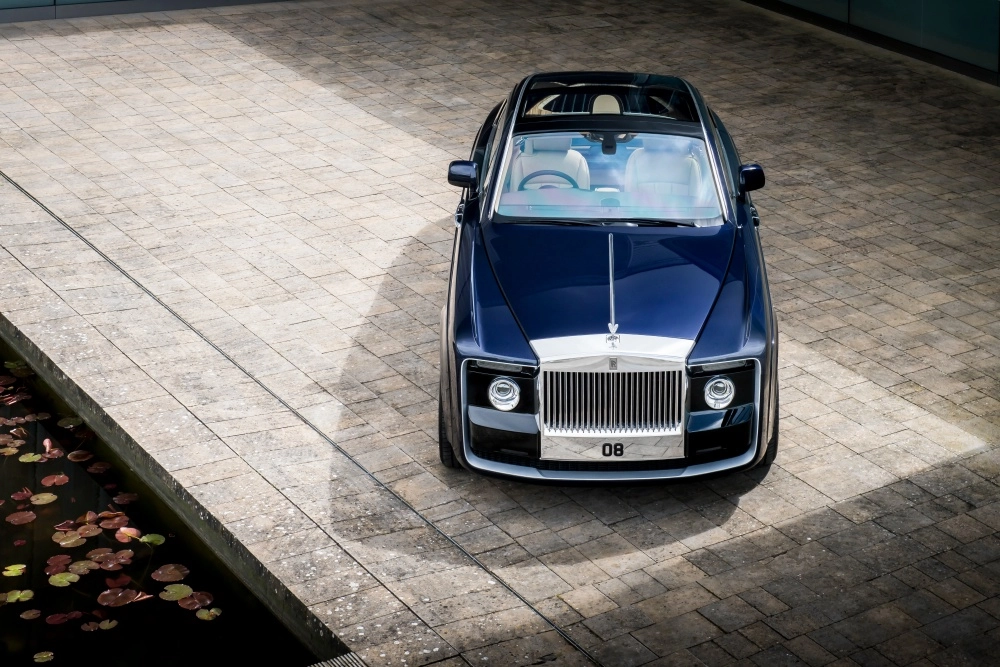 Koyu lacivert Rolls-Royce Sweptail, lüks ve limitli üretim bir otomobil olarak, yukarıdan çekilen açısıyla taş döşeli bir alanda park halinde dururken, sol tarafındaki nilüfer yapraklı su havuzuyla birlikte zarif bir şekilde görüntüleniyor ve dünyanın en pahalı otomobilleri listesindeki yerini vurguluyor.
