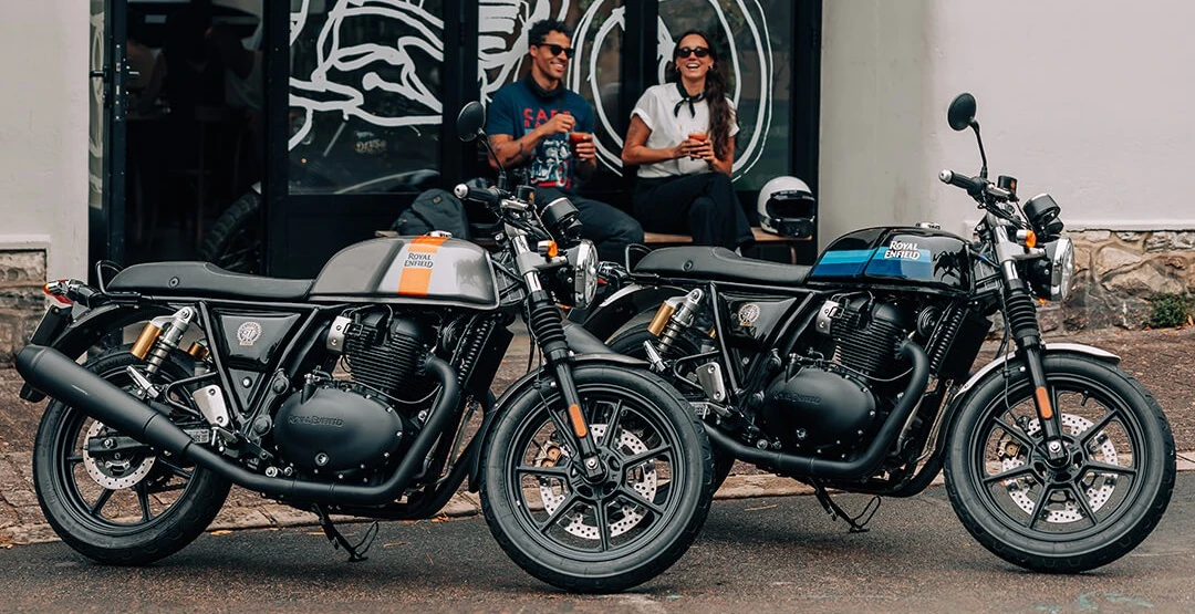 İki Royal Enfield Continental GT 650 motosiklet bir kafe önünde park edilmiş, gri ve turuncu şeritli olanı solda, siyah ve mavi şeritli olanı sağda dururken, arka planda bir bankta oturan iki kişi içecekleriyle keyifli sohbet ediyor.