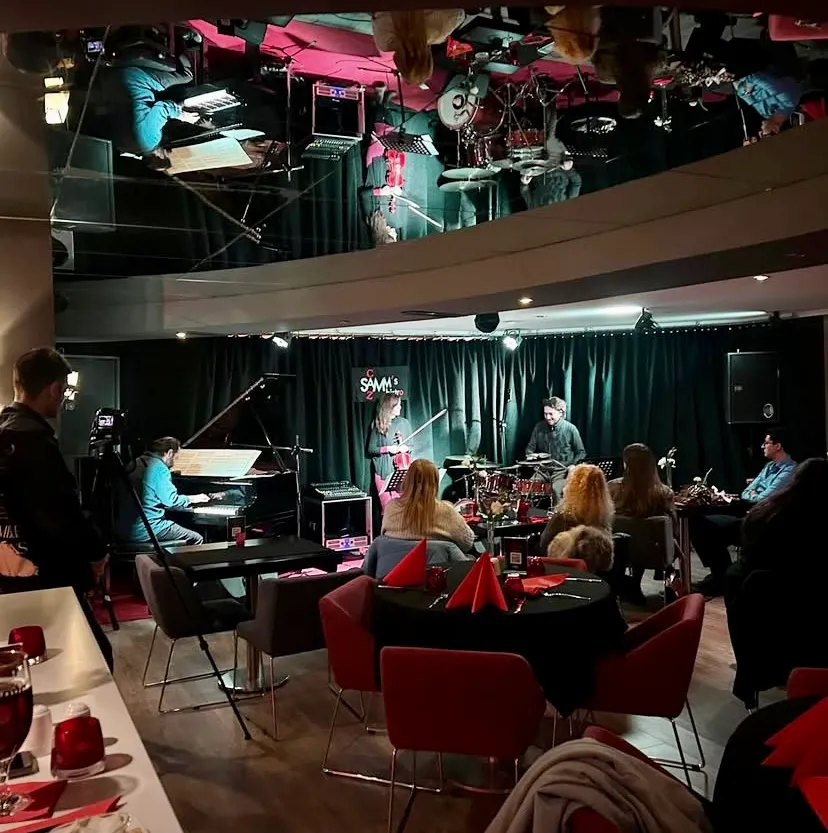 Samms Bistro Ankara'da, sahnedeki piyano, çello ve davul üçlüsünün canlı performansı sırasında, aynalı tavandan yansıyan enstrüman ve ışıklarla dolu alanda oturan misafirler samimi bir akşamın tadını çıkarıyor.