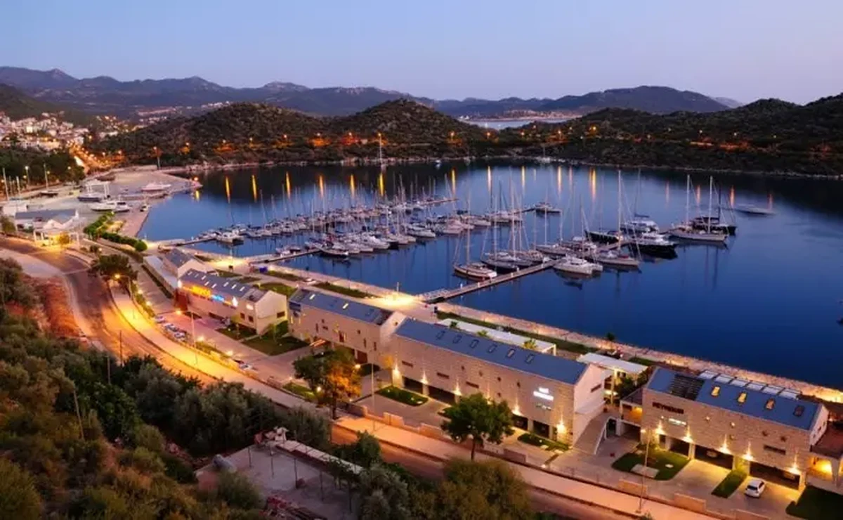 Gün batımından sonraki alacakaranlıkta, ışıkları suya yansıyan yelkenli ve yatlarla dolu Makmarin Kaş Marina, çevresindeki yeşil tepeler ve ışıklandırılmış sahil şeridiyle görülüyor.