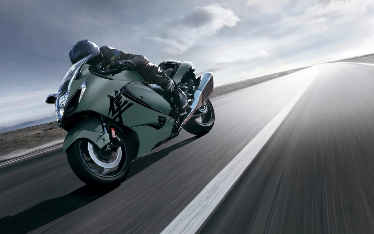 Suzuki Hayabusa yaris motosikleti ile tam donanımlı bir sürücü, hızla bir virajı alırken, gri-yeşil motosikletin detayları ve sürücünün yola eğildiği an, bulutlu bir gökyüzü altında hızın etkileyici dinamiklerini gösteriyor.