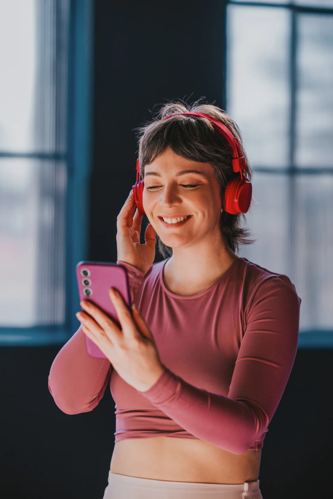 Kırmızı kulaklığıyla akıllı telefonundan müzik dinleyen mutlu genç kadın, teknoloji hediyelerinin getirdiği kişisel keyfi ve memnuniyeti yansıtıyor.