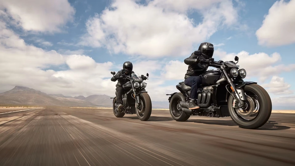 Kasklı ve deri ceketli iki sürücü, uzun ve geniş asfalt bir yolda güçlü Triumph Rocket 3R motosikletleri ile ilerlerken, arka planda dağlık arazi ve beyaz bulutlarla kaplı mavi gökyüzü dikkat çekiyor.