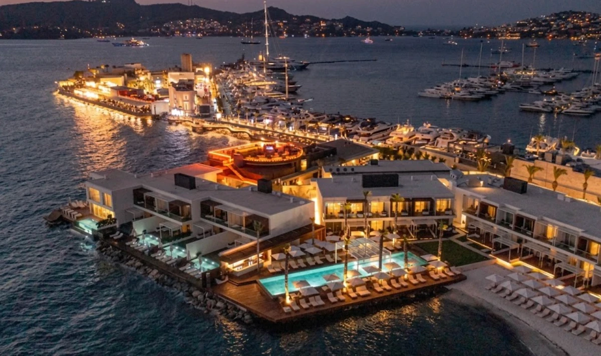 Yat turu için uygun olan akşamın lacivert tonlarında, ışıklarla parıldayan Yalıkavak Marina'nın havadan çekilmiş bu görüntüsünde, lüks yatlar ve yelkenlilerin sıralandığı modern iskeleler, hareketli restoran ve eğlence mekanları, ayrıca konforlu bir tekne tatili deneyimi sunan havuzlu otel kompleksi ve özel plaj alanı dikkat çekiyor; bu, yat turu için Türkiye'nin en seçkin marinalarından birini yansıtıyor.