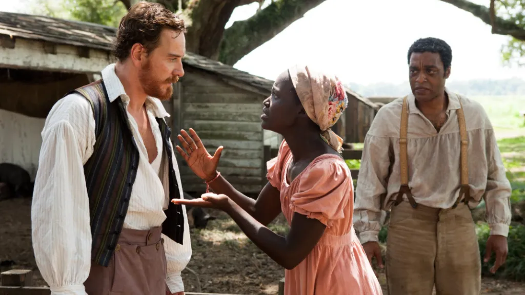 12 Yıllık Esaret (12 Years a Slave) filminden bir sahne: Genç bir kadın karakter duygusal bir şekilde konuşurken ellerini kaldırarak beyaz bir adama karşı geliyor; arka planda başka bir erkek karakter endişeyle durumu izliyor. Kırsal bir çiftlik ortamı ve eski ahşap yapılar sahneye eşlik ediyor.