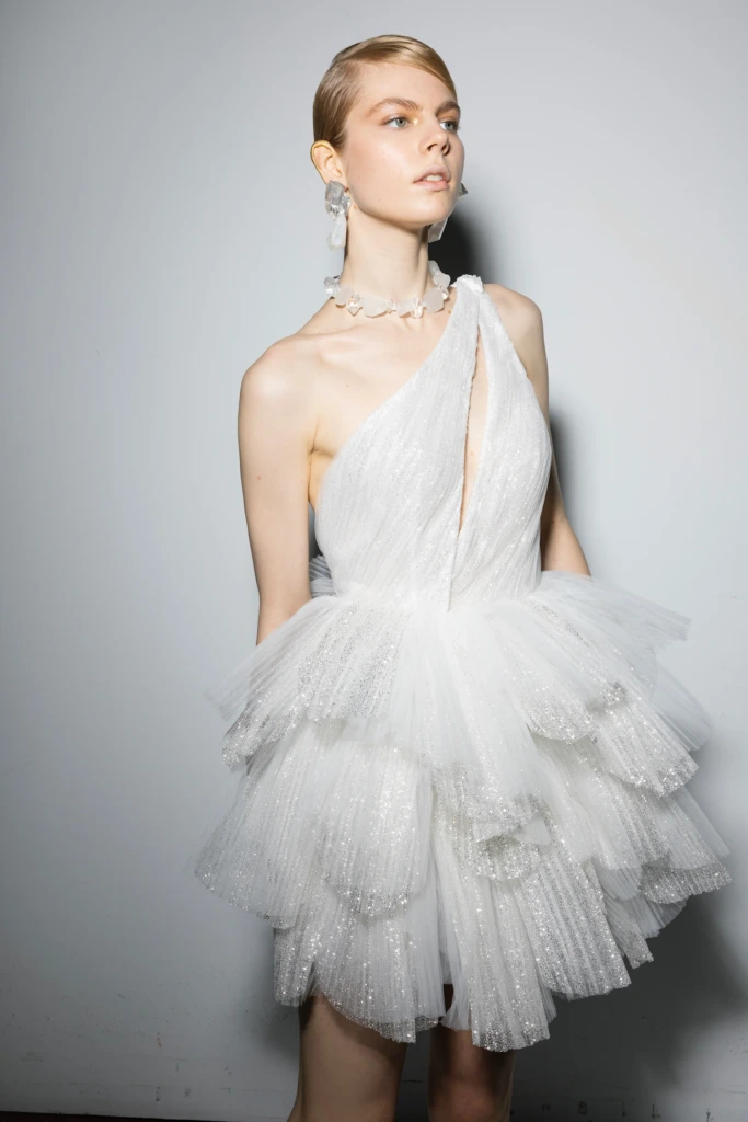 Asimetrik yakalı, derin yırtmaç detaylı ve kat kat tüllerle tasarlanmış, kısa beyaz gelinlik modeli. Işıltılı kumaşıyla dikkat çeken bu modern tasarım, avangart bir bridal parti görünümü sunuyor.