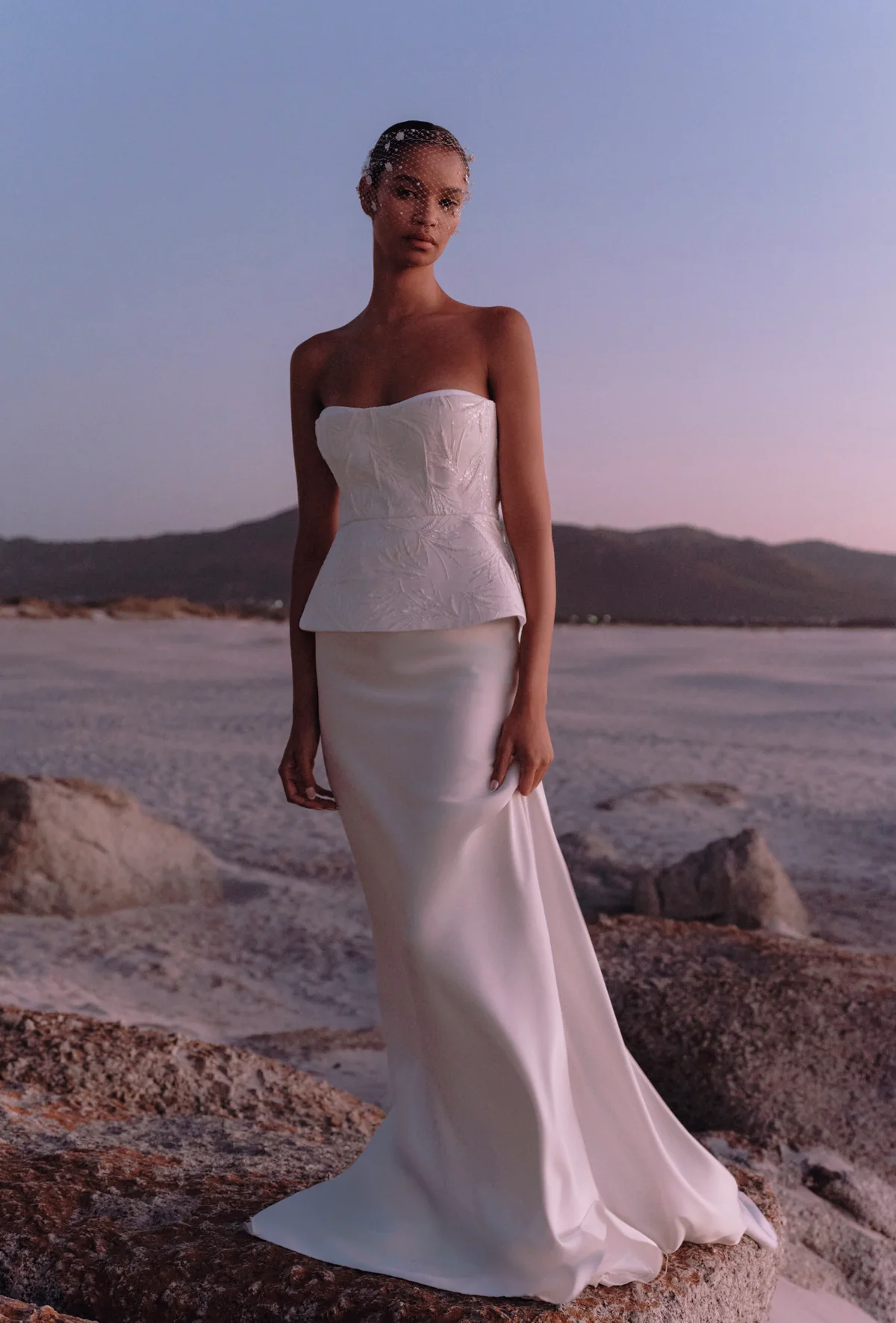 Gün batımının yumuşak renkleriyle aydınlanan geniş, açık bir alanda, dantel işlemeli straplez peplum korsajlı ve ipeksi dökümlü eteğiyle modern bir balik gelinlik modeli giymiş zarif bir kadın, küçük bir kuş kafesi duvakla poz veriyor.