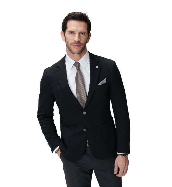 Koyu renk blazer ceket, beyaz gömlek, açık kahverengi kravat ve koyu gri pantolon giymiş, sol elini cebine atmış, lapel pini ve cep mendiliyle detaylandırılmış şık bir kombini sergileyen, bileklikleriyle modern bir hava katan, kameraya güvenle bakan erkek, makaledeki parfüm ve stil önerileri için ideal bir duruş sunuyor.