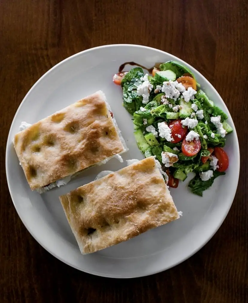 Beyaz bir tabağın üzerinde, üzeri hafif kızarmış ve karakteristik çukurlara sahip focaccia ekmeği arasına peynirli bir dolgu konularak hazırlanmış ikiye bölünmüş sandviç ile yanında bol yeşillik, kiraz domates, salatalık ve beyaz peynir içeren taze bir salata sunuluyor.