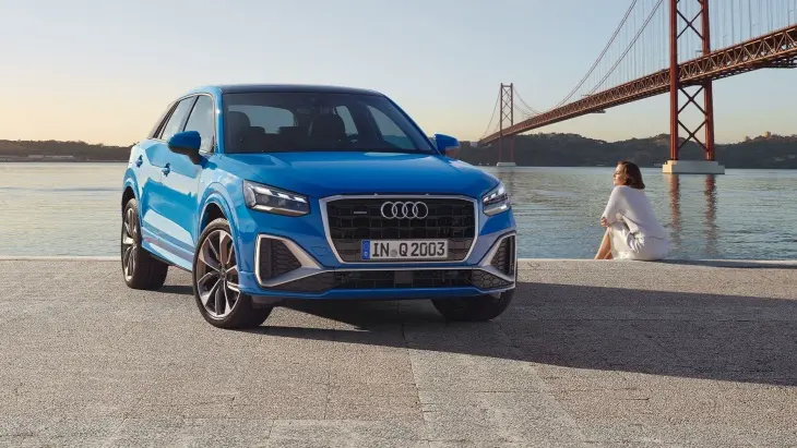 Parlak mavi renkte bir Audi Q2 model araba, denize bakan bir rıhtımın üzerinde duruyor. Arka planda bir köprü ve denize karşı oturmuş beyaz elbiseli bir kadın görülüyor.