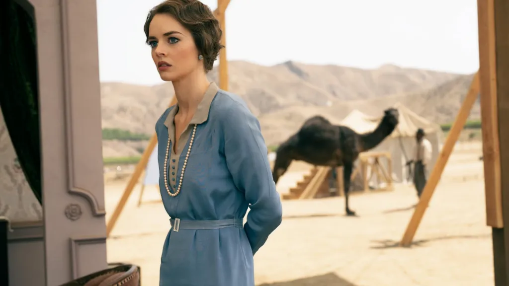 Babylon (Babil) filminden bir sahne: Çölün ortasında kurulmuş bir film setinde, 1920'ler tarzı mavi saten bir elbise giymiş, boynunda inci kolye olan genç bir kadın endişeli bir ifadeyle ileriye bakıyor. Arka planda bir deve, çadırlar ve dönemin kıyafetlerini giymiş bir adam bulunuyor. Kadının bulunduğu yer, setin kenarında bir çadırın girişine yakın bir noktada. Atmosfer, eski Hollywood'un altın çağını ve egzotik film prodüksiyonlarını yansıtıyor.