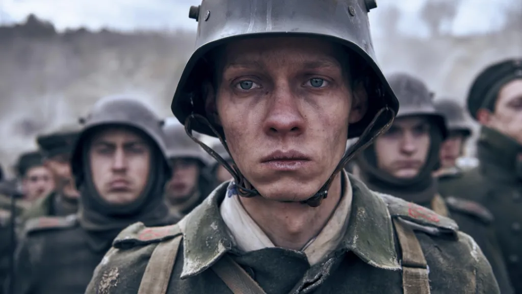 Batı Cephesinde Yeni Bir Şey Yok (All Quiet on the Western Front) filminden çarpıcı bir sahne: Genç bir asker çamur içindeki yüzüyle kameraya donuk ve yorgun bakarken, arkasında sıralanmış diğer askerler savaşın ağırlığını taşıyan ifadelerle duruyor. Gri ve puslu hava, sahnenin karanlık atmosferini pekiştiriyor.