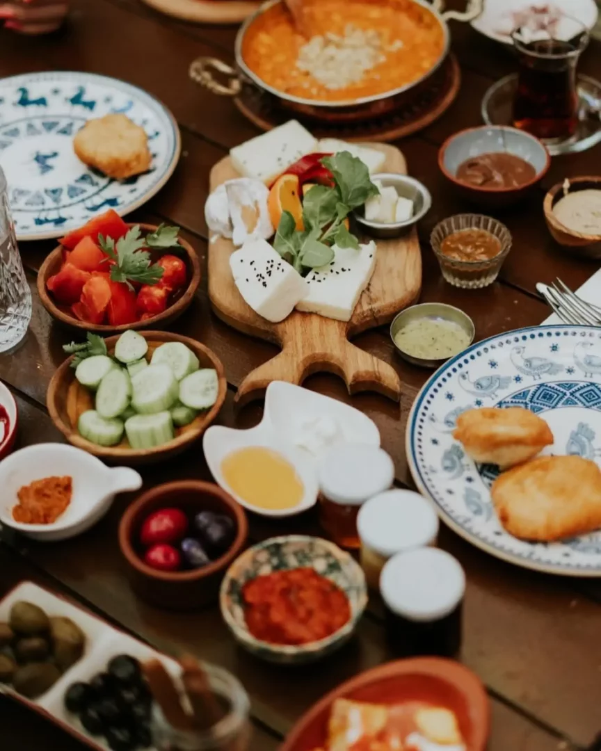 Masada, peynir çeşitleri, dilimlenmiş domates ve salatalık, menemen benzeri bir tava yemeği, zeytinler, pişi, bal, reçel, tahin-pekmez ve çay gibi birçok lezzetli yiyecekle donatılmış zengin bir Türk kahvaltısı sofrası göze çarpıyor; bu cömert kahvaltı, bir Breakfast Club deneyimini anımsatan samimi ve keyifli bir buluşmayı çağrıştırıyor.