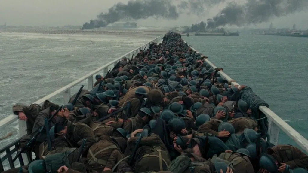 Dunkirk filminden yoğun bir savaş sahnesi: Askerlerle dolu bir iskelede, yüzlerce asker yere eğilmiş halde bombardımandan korunmaya çalışıyor. Arka planda dumanlar yükseliyor ve kasvetli bir hava hâkim. Sahne, Dunkirk Tahliyesi’nin kaotik ve umutsuz atmosferini yansıtıyor.