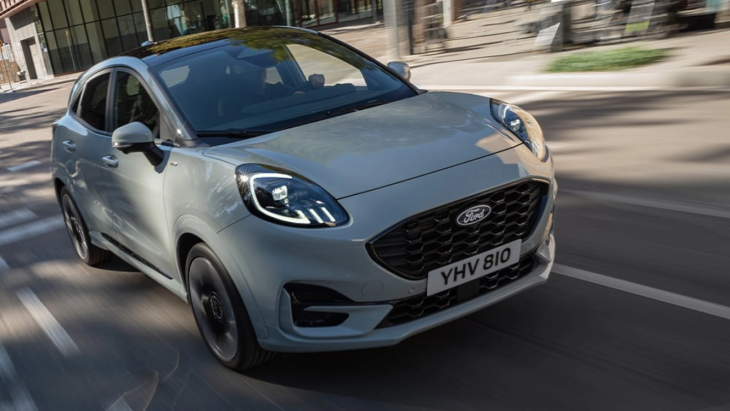 Gri renkte bir Ford Puma model araba, şehirde yolda ilerlerken çekilmiş. Aracın dinamik tasarımı ve far detayları ön planda, arka planda ise modern binalar görülüyor.