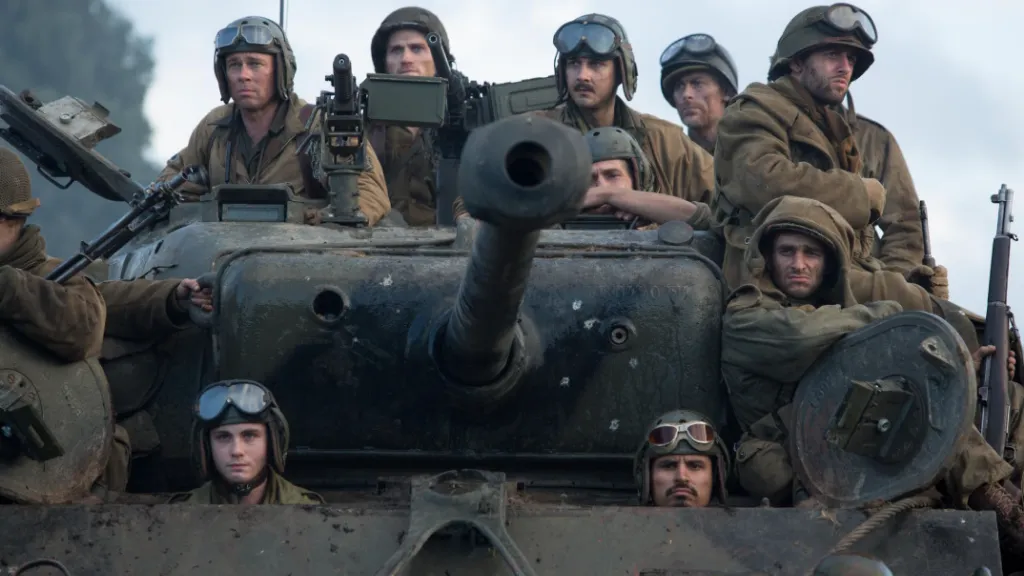 Fury filminden bir sahne: II. Dünya Savaşı'nda geçen bu karede, bir grup Amerikan askeri zırhlı bir tankın üzerinde toplanmış şekilde ileriye bakıyor. Askerlerin yüzleri yorgunluk, kararlılık ve gerginlik ifadeleriyle dolu. Tankın namlusu doğrudan kameraya yöneltilmiş. Sahne, savaşın sert gerçekliğini ve birlik ruhunu yansıtıyor.