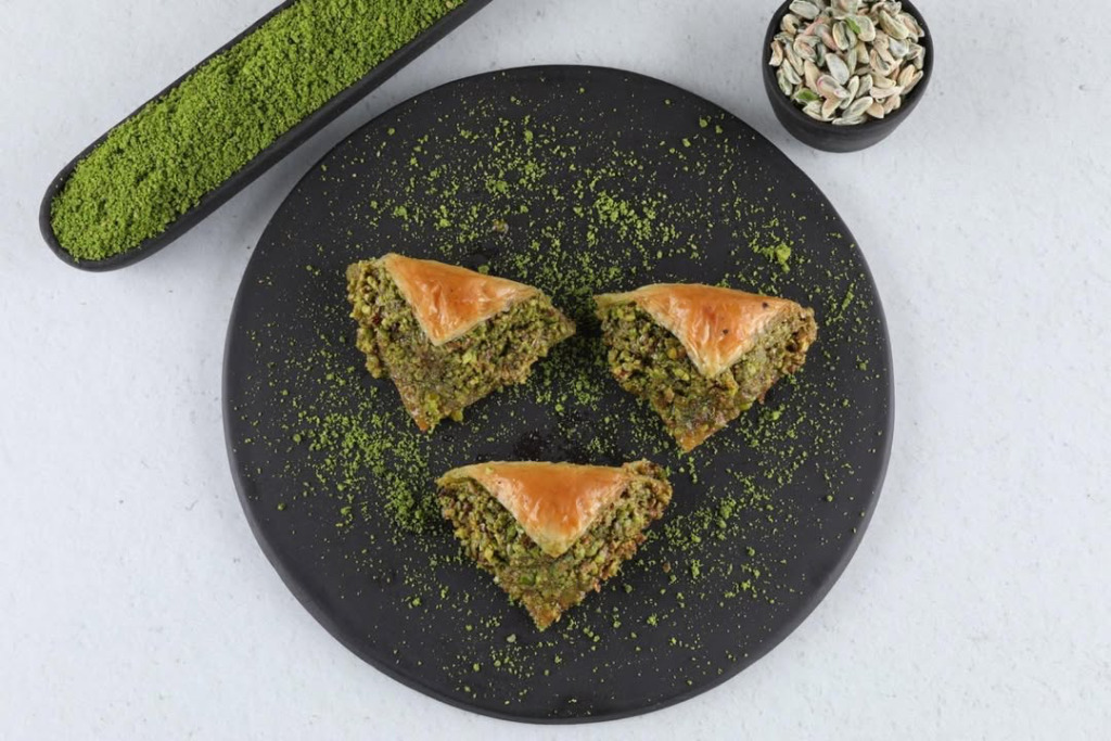 Siyah yuvarlak tabakta sunulan üç adet bol fıstıklı üçgen dilim Hamido Baklava. Çıtır yufkaların üzeri parlak yeşil antep fıstığı tozuyla süslenirken, arka plandaki toz ve tane fıstık kaseleri bu eşsiz Türk lezzetinin zenginliğini