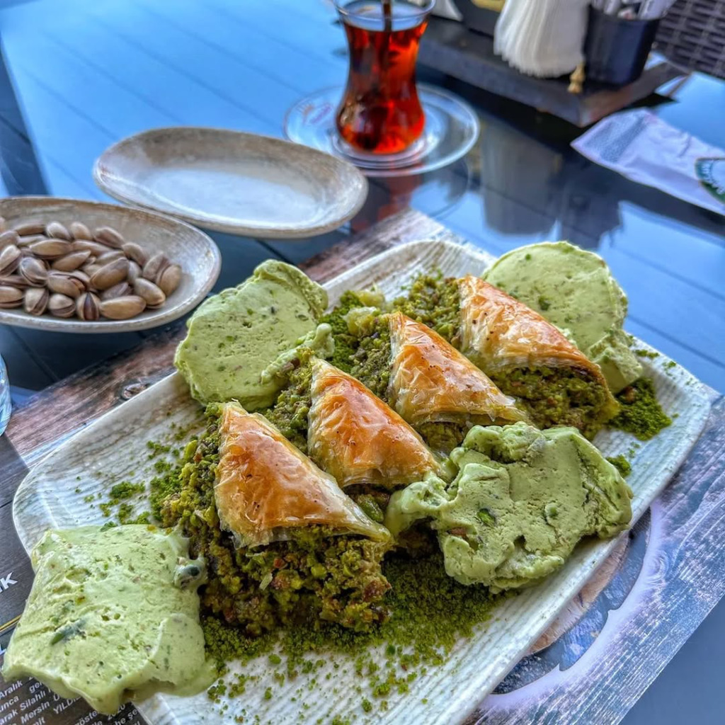 Hamido Baklava'nın bol fıstıklı dondurma ve öğütülmüş fıstık yatağında sunulan çıtır midye baklavaları iştah kabartıyor. Arka planda ince belli bardakta Türk çayı ve bütün Antep fıstıkları bu eşsiz geleneksel lezzeti tamamlıyor.