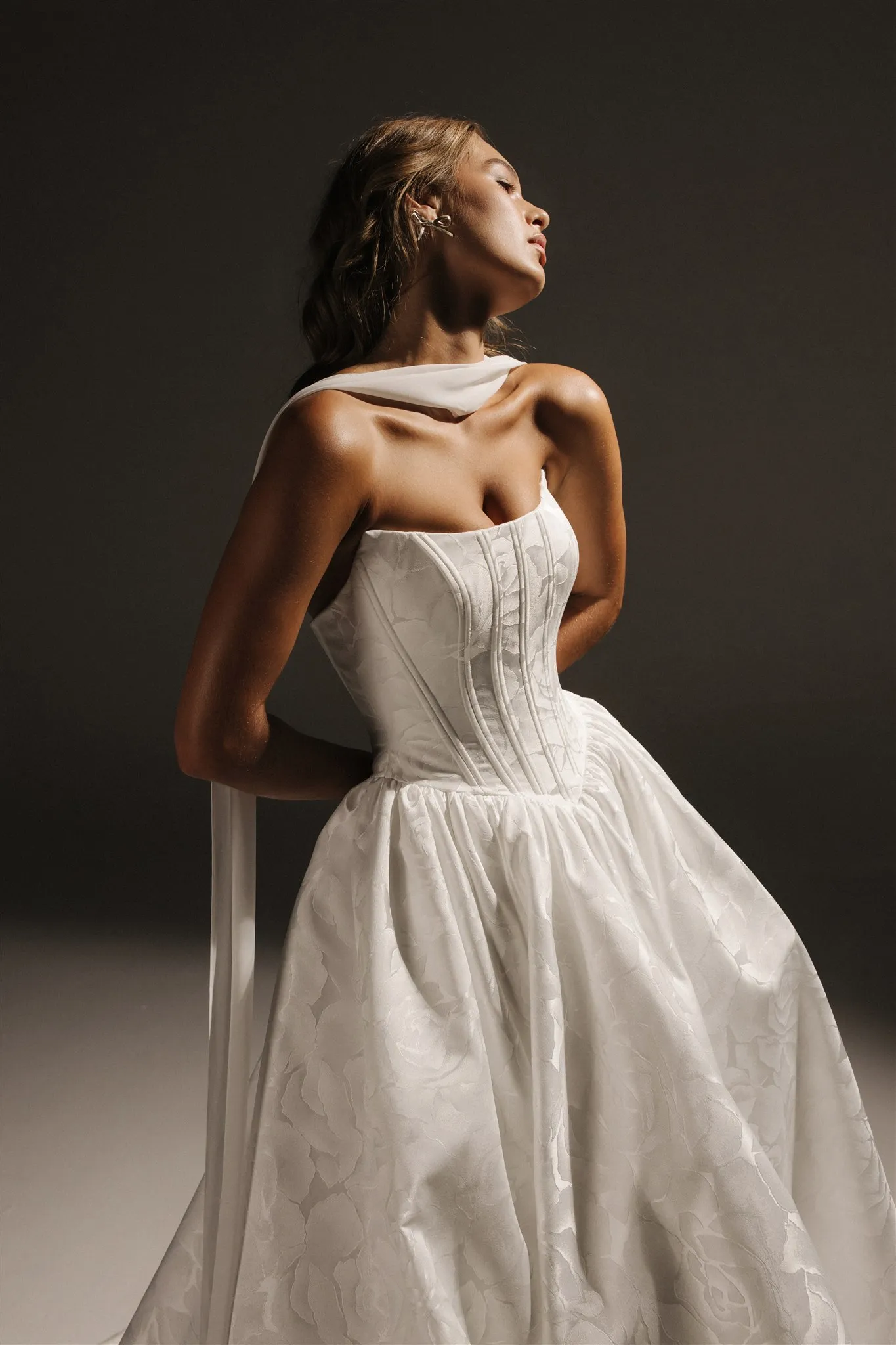 Romantik zarafetin modern yorumunu sunan Hera Couture gelinlik, dantel görünümlü jakarlı kumaşı ve yapılandırılmış korse detayıyla nostaljik ama güçlü bir duruş sergiliyor. Omuzdan süzülen ince şal detayı ise görünüme zarif bir şiirsellik katıyor.