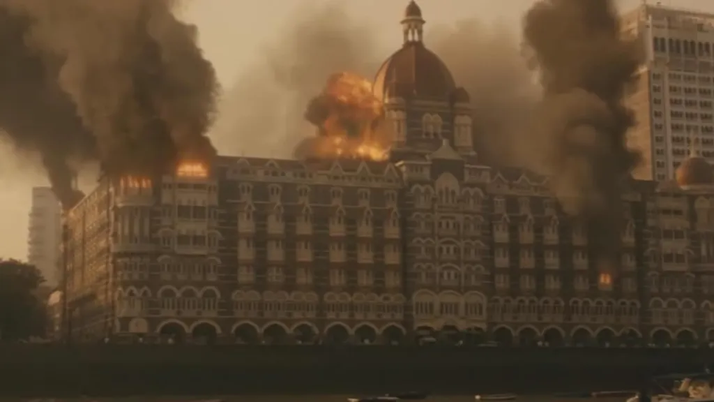 Hotel Mumbai filminden bir sahne: Lüks bir otel binası alevler içinde yanarken kalın siyah dumanlar gökyüzüne yükseliyor. Patlamalar binanın farklı bölümlerini sarmış durumda. Sahne, 2008 Mumbai terör saldırılarının dehşet verici boyutunu ve yıkıcılığını dramatik bir şekilde yansıtıyor.