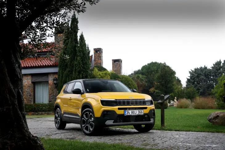Sarı renkte bir Jeep Avenger model araba, lüks bir evin taş döşeli otoparkında duruyor. Aracın arka planında bahçesi ve mimari detayları görünen bir villa yer alıyor.