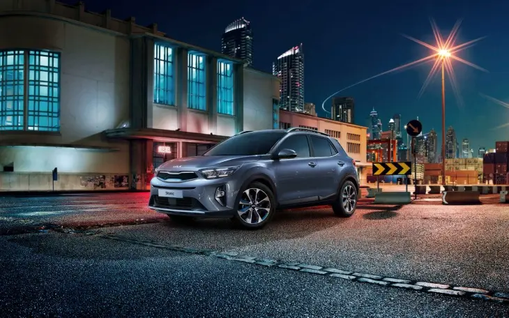 2024 model gri Kia Stonic, gece vakti şehirde aydınlatılmış binaların ve sokak lambalarının önünde park halinde. Fotoğraf, aracın modern ve dinamik tasarımını, keskin hatlarını ve şık LED farlarını vurguluyor. SUV model, şehir içi kullanıma uygun boyutlarıyla dikkat çekiyor.