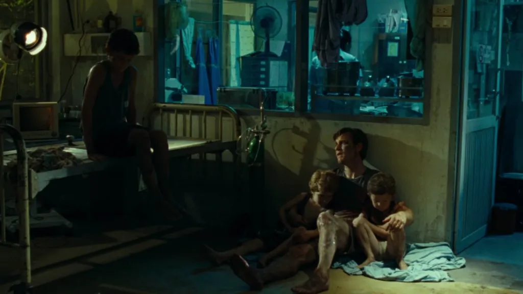 The Impossible (Kıyamet Günü) filminden bir sahne: Tayland’daki tsunami felaketinden sonra hastaneye sığınmış bir baba ve çocukları, yerde birbirine sarılmış şekilde oturuyor. Arka planda yarı karanlık bir hastane odası, eski yataklar ve tıbbi ekipmanlar görülüyor. Sahne, felaket sonrası yaşanan travmayı ve aile bağlarının gücünü yansıtıyor.