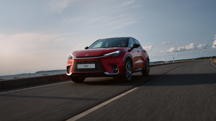 Kırmızı renkte bir Lexus LBX model araba, sahil yolu gibi görünen bir yolda ilerliyor. Arka planda deniz, dağlar ve güneşli bir gökyüzü manzarası bulunuyor.
