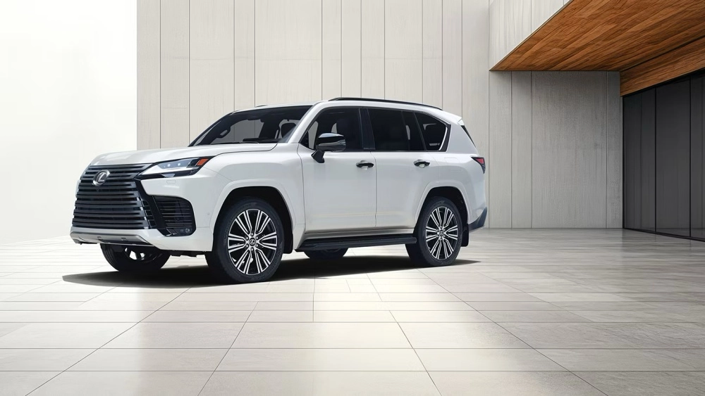 Beyaz renkli, belirgin ızgarası ve dinamik jantlarıyla dikkat çeken modern bir Lexus LX 700 lüks SUV, açık renkli fayans zemin üzerinde, sade ve çağdaş mimariye sahip bir yapının önünde sol ön köşesinden çekilmiş bir açıyla aracın etkileyici dış mekan tasarımını sergiliyor.