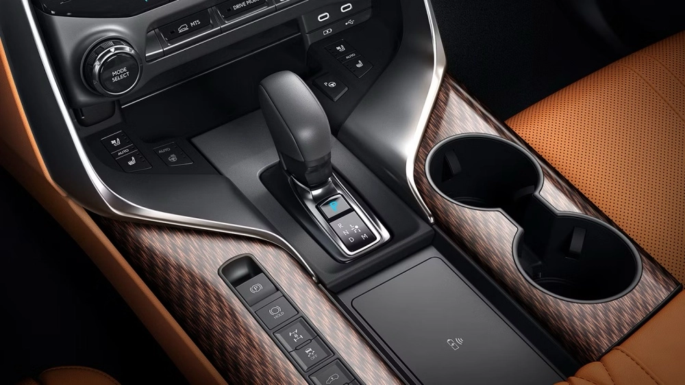 Bir Lexus LX 700 lüks SUV'nin iç mekanından detaylı bir görünüm; ahşap desenli kaplamalara sahip orta konsolu, otomatik vites kolunu, sürüş modu seçici düğmesini, ısıtmalı koltuk ve direksiyon kontrollerini, kablosuz şarj pedi ve bardak tutucularını gösteriyor.