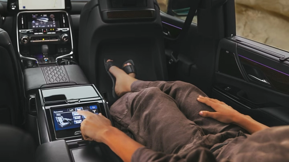 Lexus LX 700 model bir lüks SUVun arka makam koltuğunda, kahverengi rahat kıyafetler giymiş bir kişi, bacaklarını uzatmış bir şekilde dinlenirken koltuk masaj ayarlarını kontrol eden dokunmatik ekranı parmağıyla kullanmakta ve aracın modern iç mekan tasarımı ile konforu ön plana çıkmaktadır.