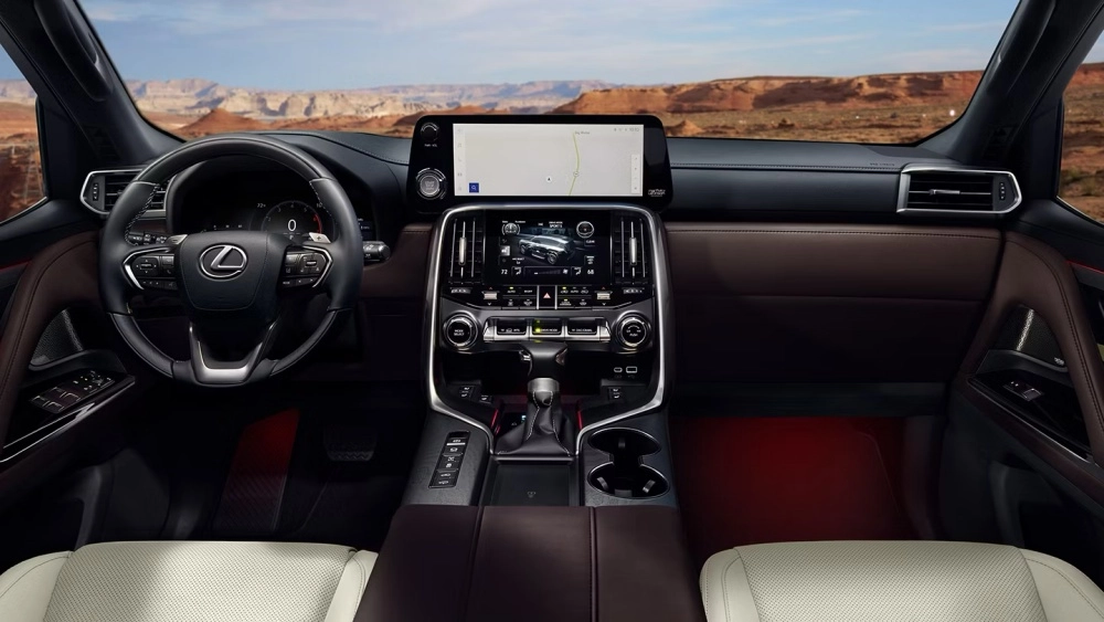 Lexus LX 700 lüks SUV'un iç mekan tasarımını ve modern ön konsolunu gösteren bu görüntüde, dijital gösterge paneli, haritalı üst ekran, araç bilgilerini sunan alt ekran, kahverengi deri kaplamalar, beyaz delikli koltuklar ve ön camdan görünen dağlık çöl manzarası öne çıkıyor.