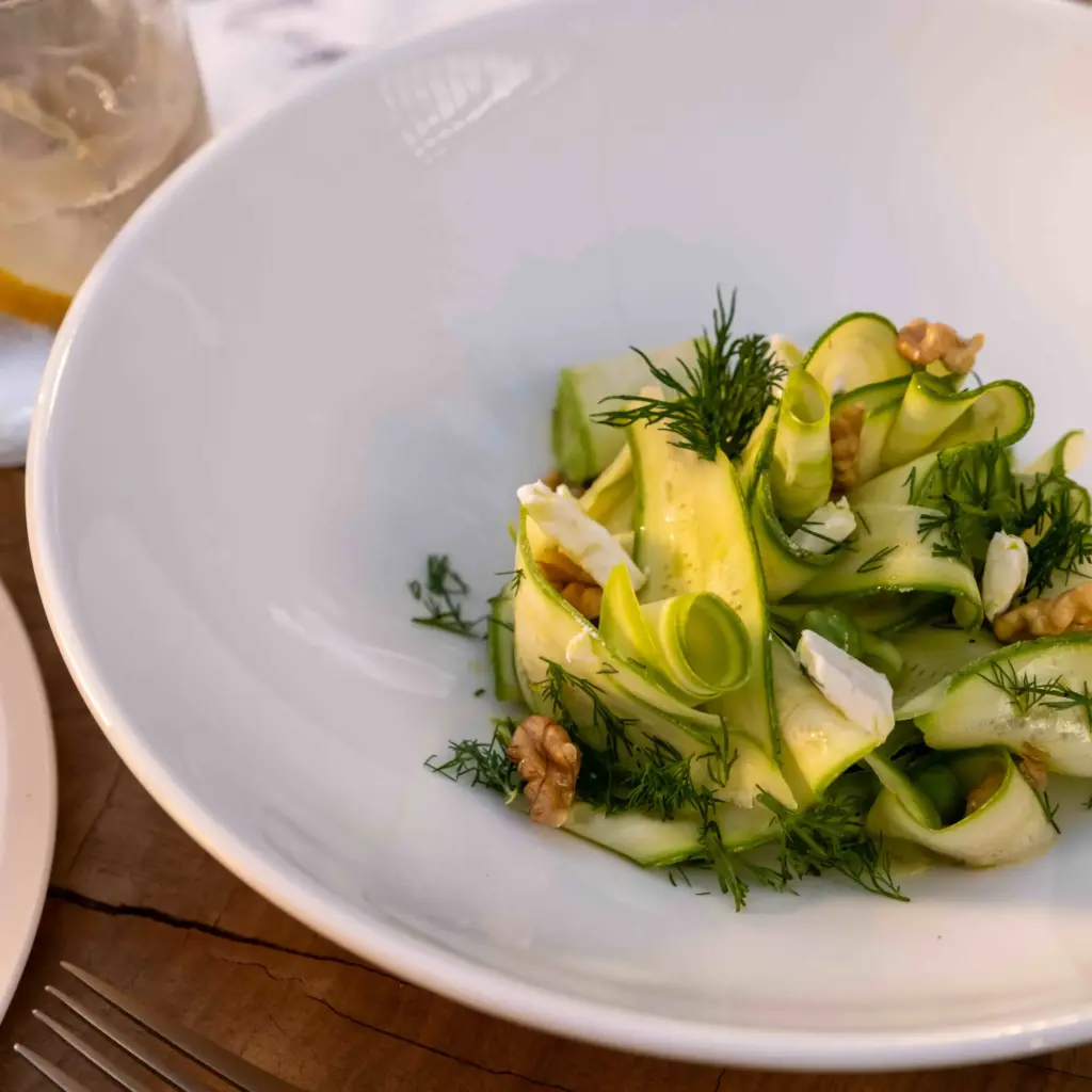 Beyaz derin bir kasede sunulan, ince dilimlenmiş yeşil kabak şeritleri, taze dereotu, parçalanmış ceviz ve beyaz peynirlerle zenginleştirilmiş ferahlatıcı bir salata, Bodrum restoranlarının modern Ege mutfağına sunduğu lezzetli başlangıçları yansıtıyor.