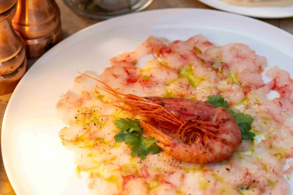 Bodrum’un en iyi restoranları arasında öne çıkan bir mekanda sunulan, zeytinyağı ve taze çekilmiş karabiberle lezzetlendirilmiş ince dilimlenmiş pembe karides carpaccio'sunun, üzerinde bütün bir kırmızı karides ve yeşil maydanoz yaprağıyla zarifçe süslenmiş iştah açıcı sunumu.