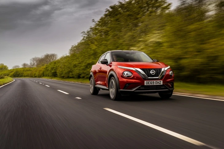 Kırmızı renkte bir Nissan Juke model araba, kırsal bir yolda ilerlerken çekilmiş. Arka planda yemyeşil ağaçlar ve tarlalar görünüyor.