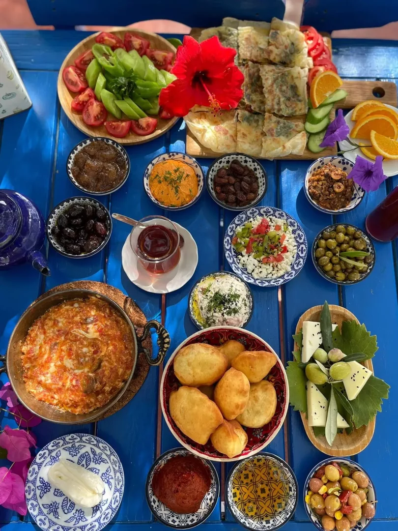 Birçok geleneksel Türk kahvaltı lezzetinin sunulduğu, menemen, pişi, gözleme, zeytin çeşitleri, peynirler, reçeller, bal ve taze sebzelerle dolu, Alaçatı kahvaltısının zenginliğini ve misafirperverliğini yansıtan mavi bir masadaki cömert serpme kahvaltı sofrası.